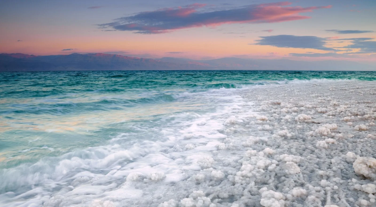 Dead Sea salt crust along the shoreline at sunset, with the escarpment on the far side of the water