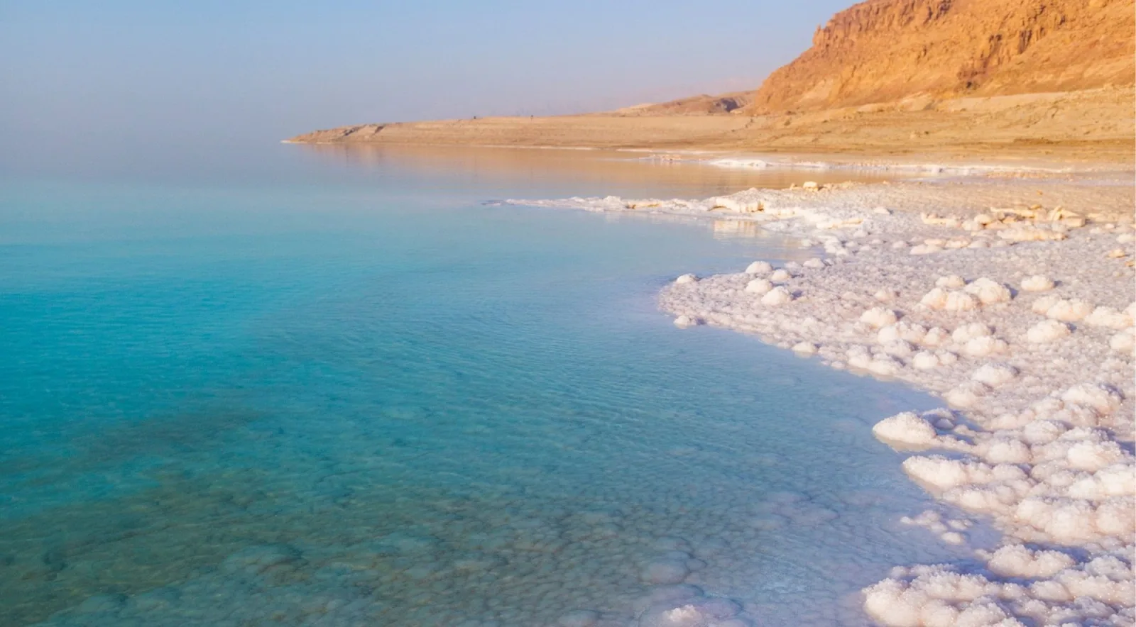 Calm turquoise Dead Sea water beside a white salt beach with the cliffs rising behind