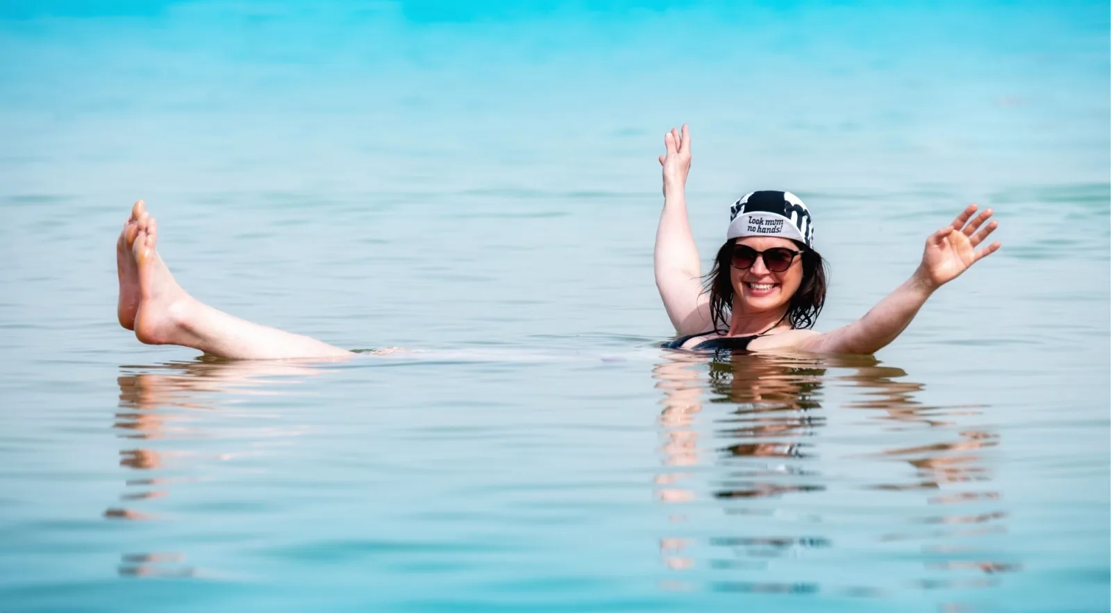 A woman floating on her back in bright turquoise Dead Sea water with both arms in the air