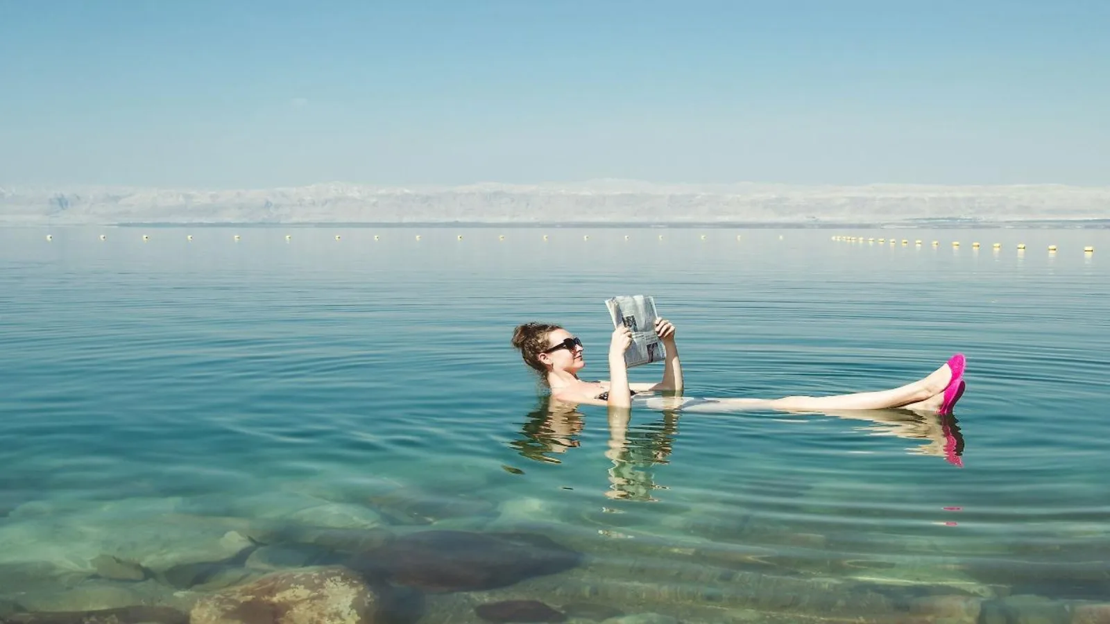 A woman floating on her back in the Dead Sea reading a newspaper, the far shore behind her