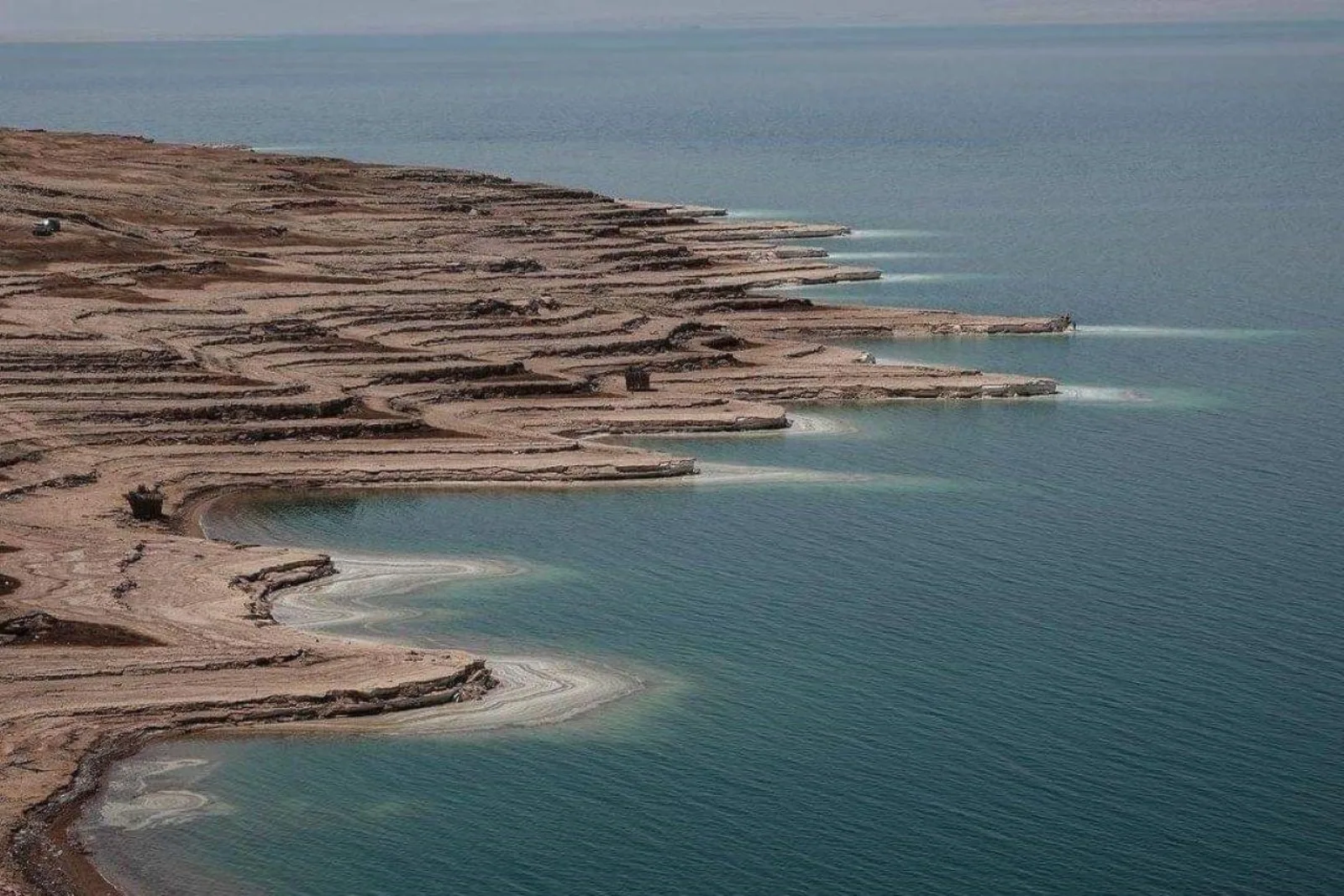 The scalloped Dead Sea shoreline from above, salt terraces cutting into green-blue water