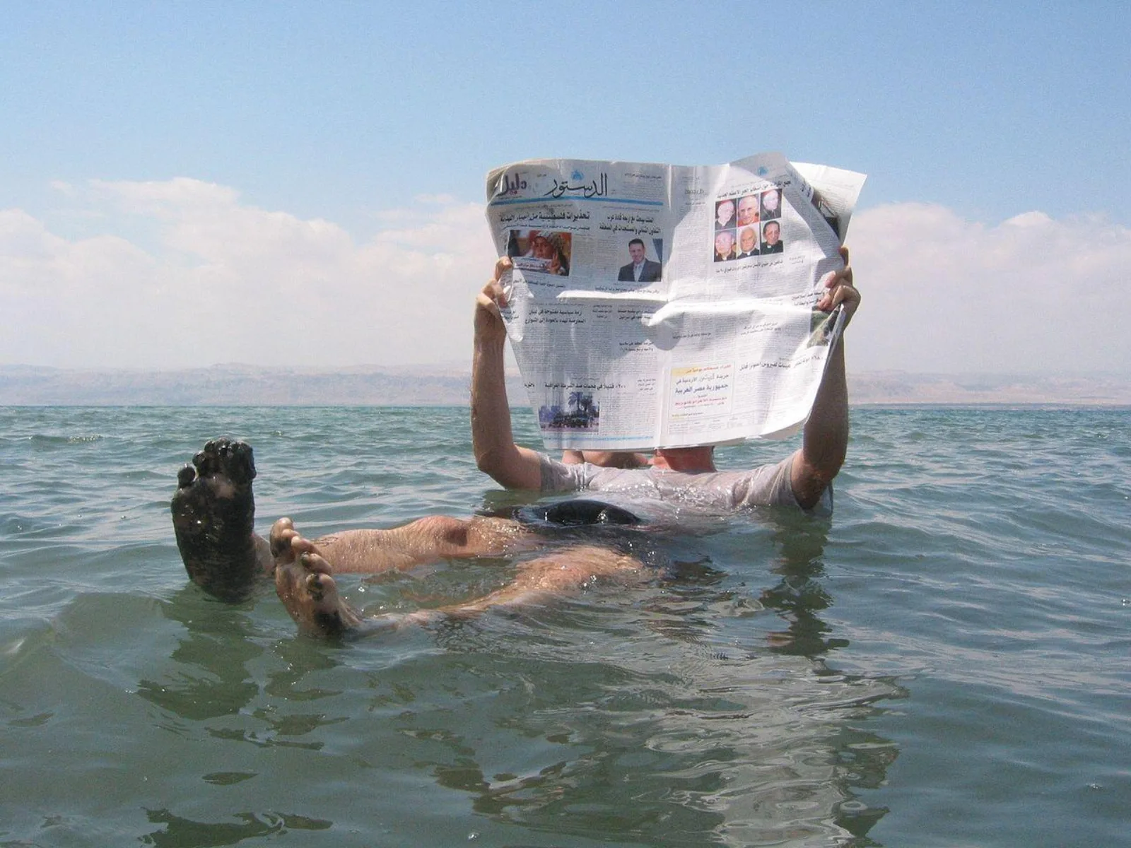 A man floating on his back in the Dead Sea reading an Arabic newspaper, feet coated in mud