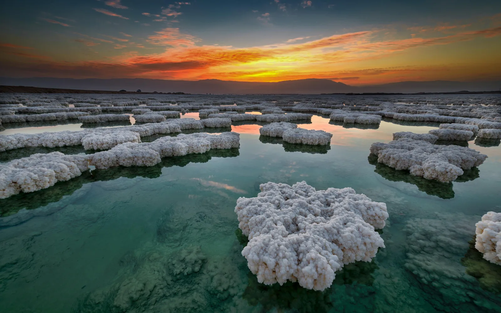 Salt formations across the Dead Sea shallows at sunset, the hills silhouetted behind