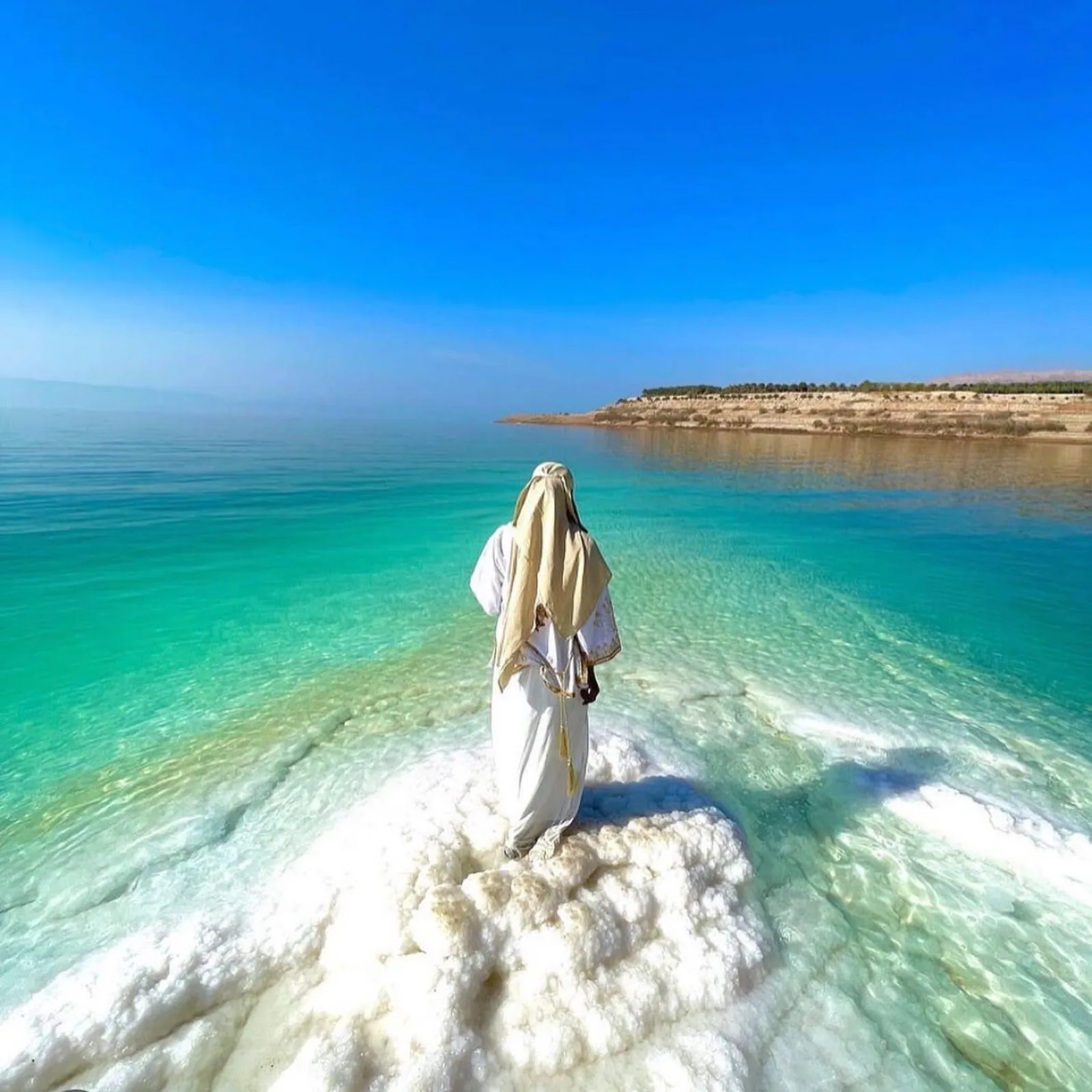 A figure in white robes standing on a salt shelf above clear turquoise Dead Sea water
